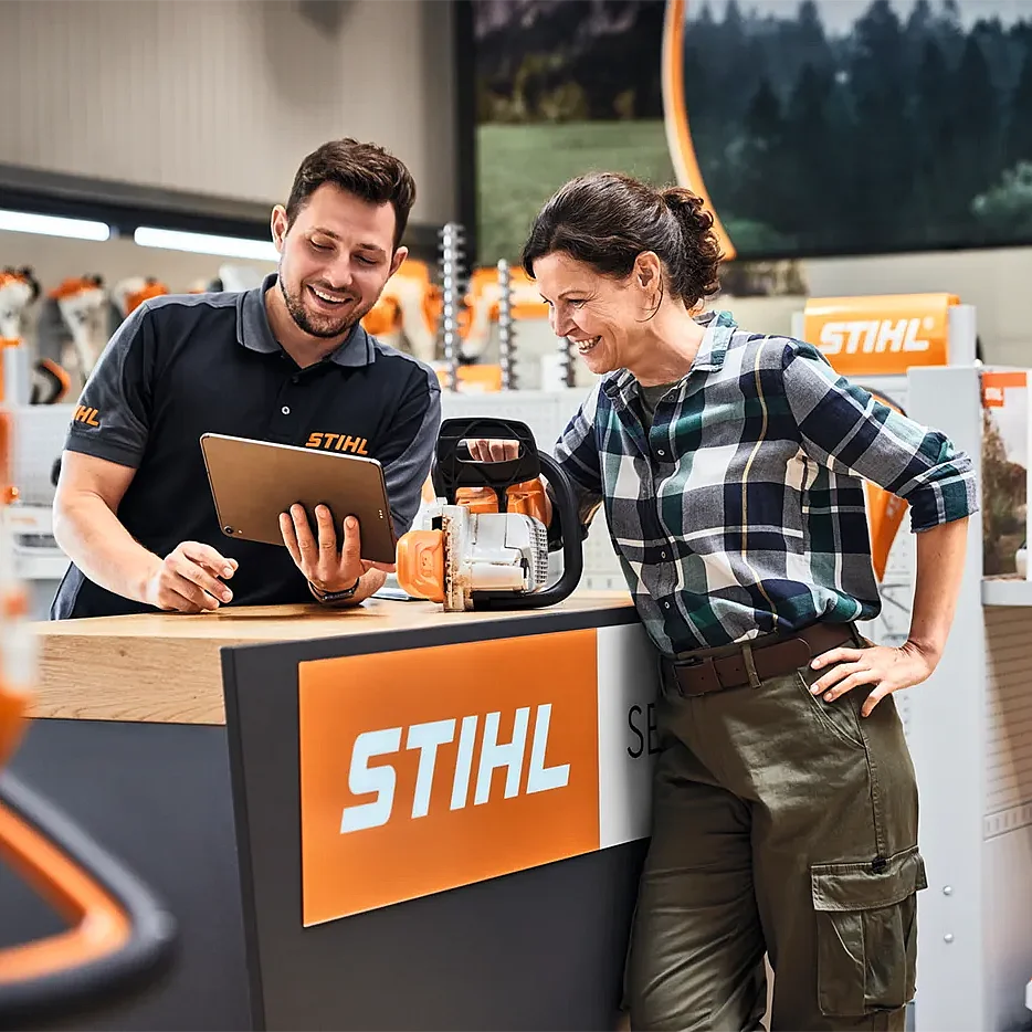 STIHL employee advising a customer at a retail counter, presenting a chainsaw and digital product information in a branded store environment.