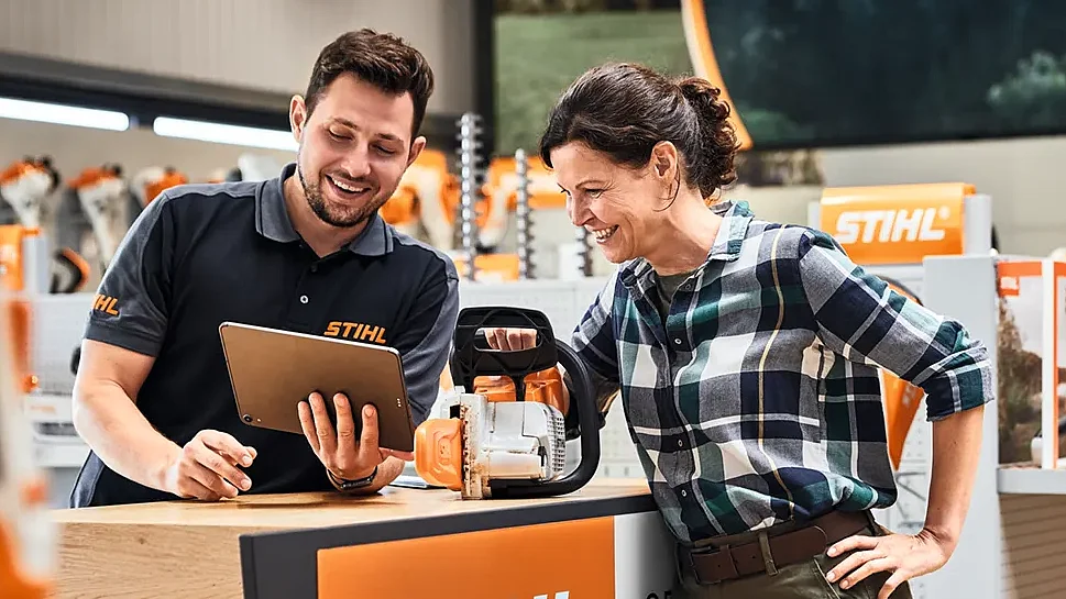 STIHL employee advising a customer at a retail counter, presenting a chainsaw and digital product information in a branded store environment.