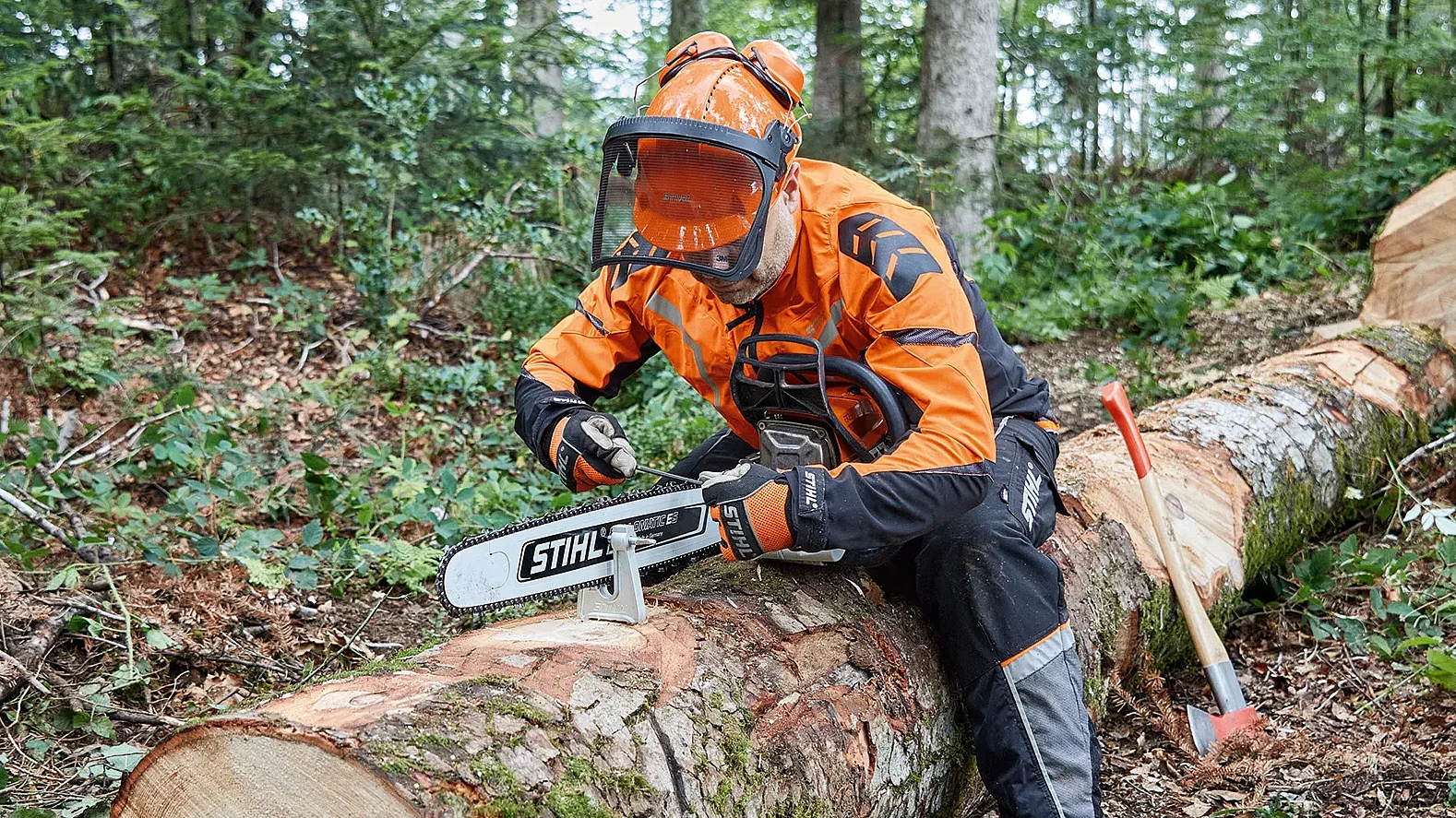 Forestry worker in protective STIHL gear sharpening a chainsaw while sitting on a felled tree trunk in a forest.