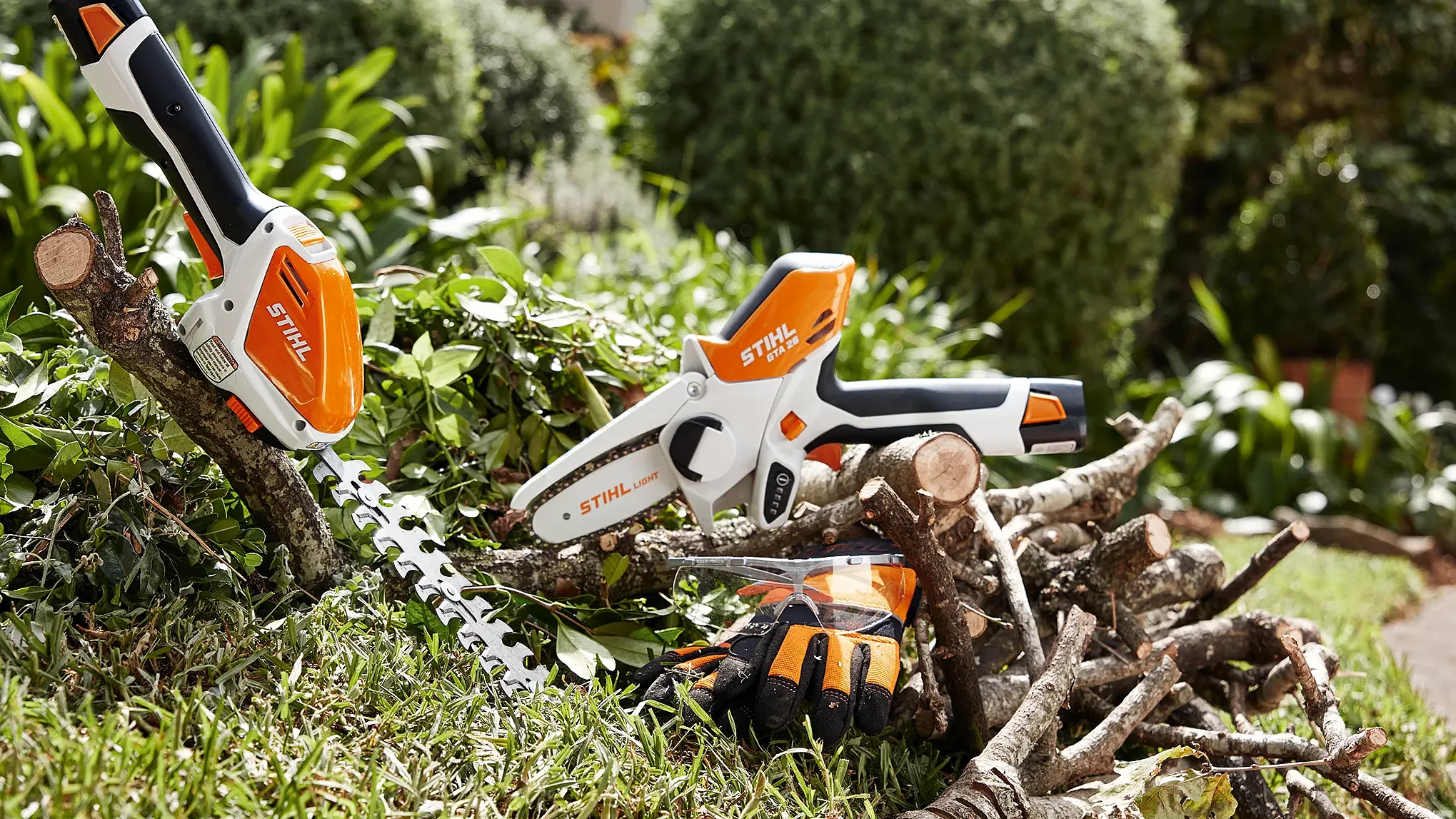 STIHL cordless hedge trimmer and compact chainsaw placed on garden branches, with gloves and a landscaped backyard in the background.
